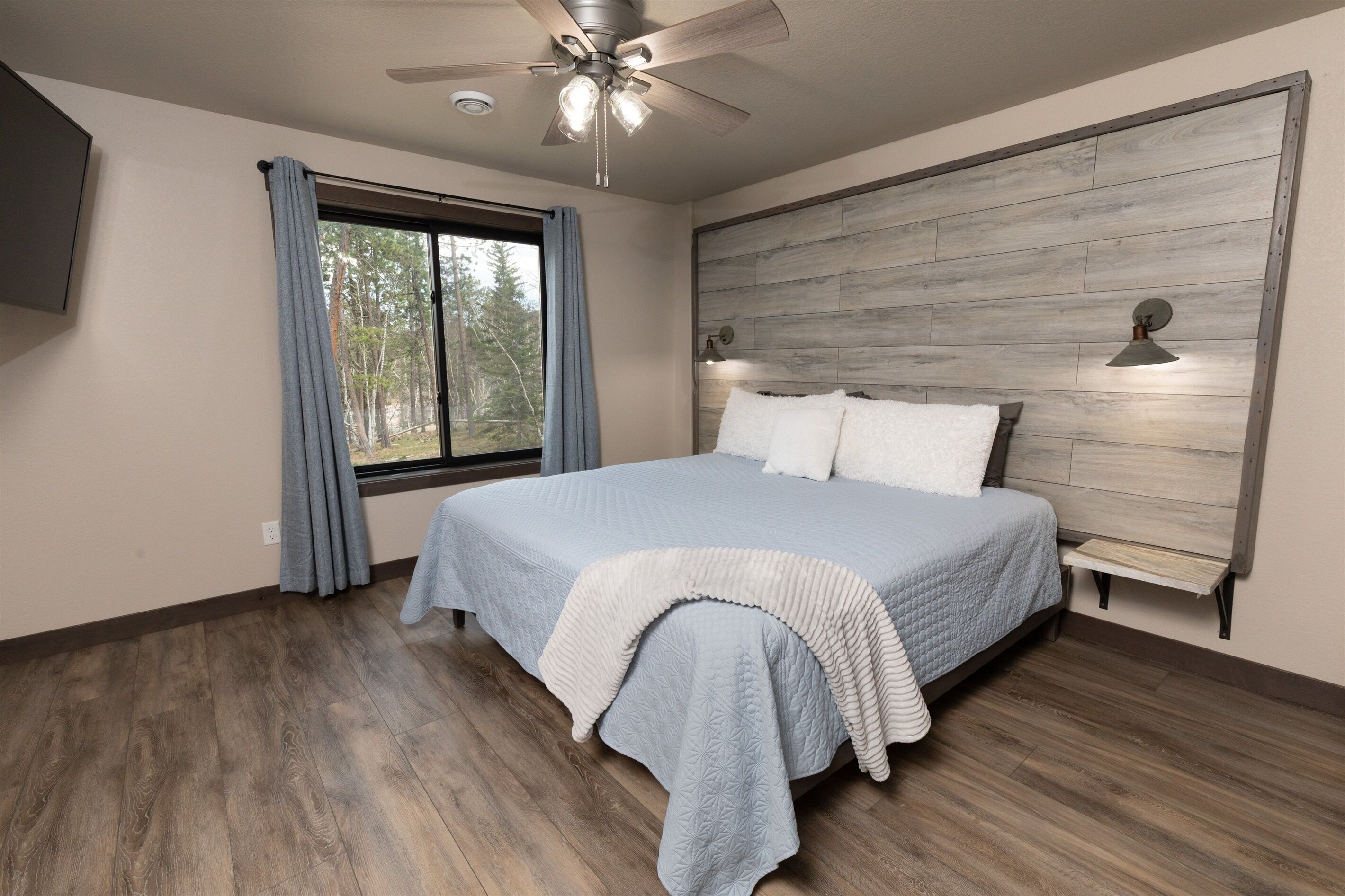 Une chambre à coucher comprenant un lit, un ventilateur de plafond, une fenêtre avec des rideaux et un luminaire fixé au mur.