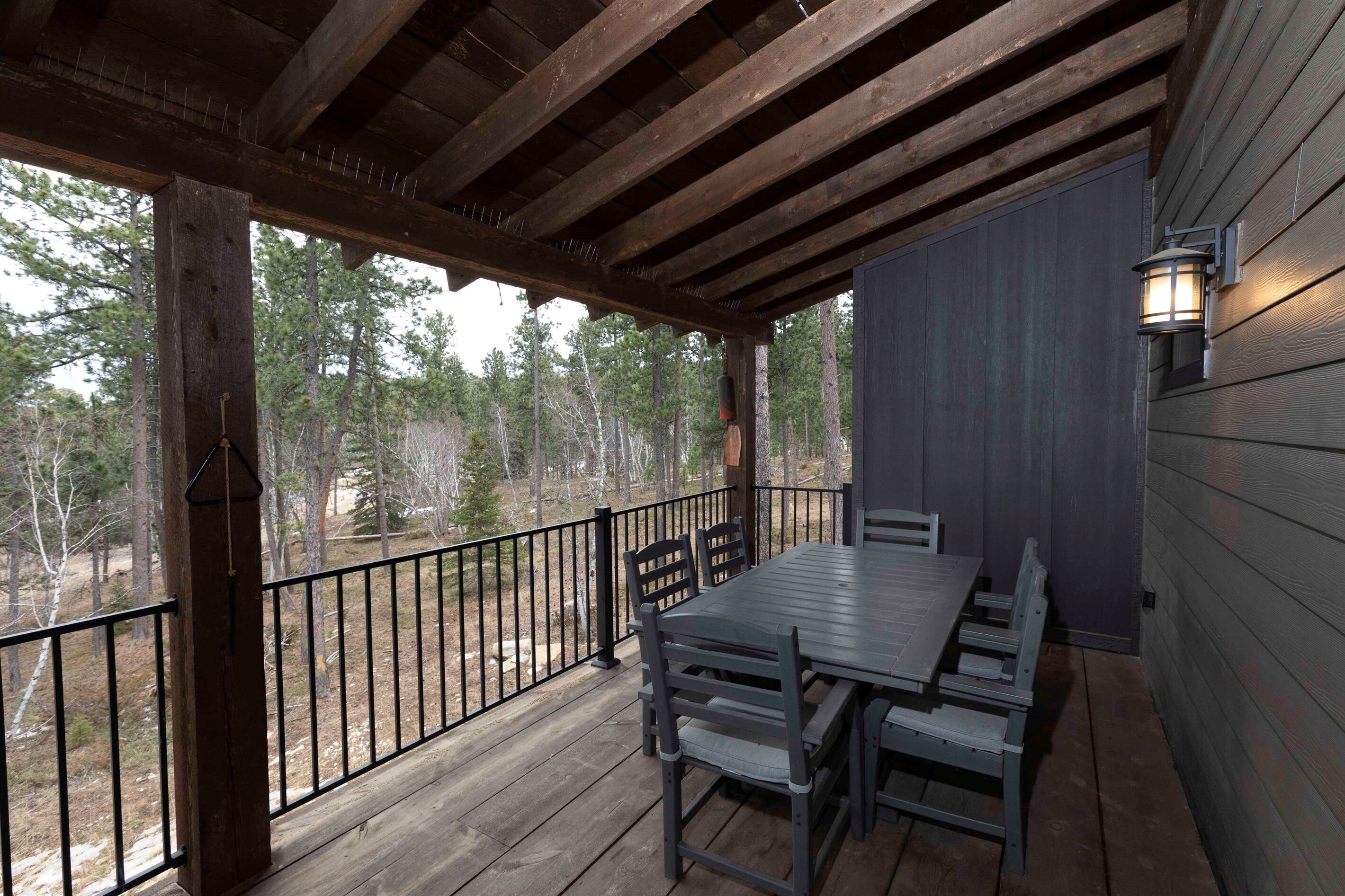 A covered outdoor area with a wooden table and chairs, a railing, and a view of trees.