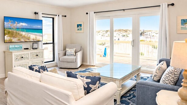 Una sala de estar con sofá, sillón y una mesa de centro. Hay un televisor en la pared, un mueble y vistas a la playa a través de las ventanas.
