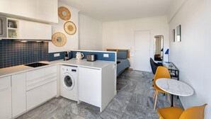Une cuisine compacte équipée d’un lave-linge, un coin repas avec une table et des chaises, et un lit à l’arrière-plan.
