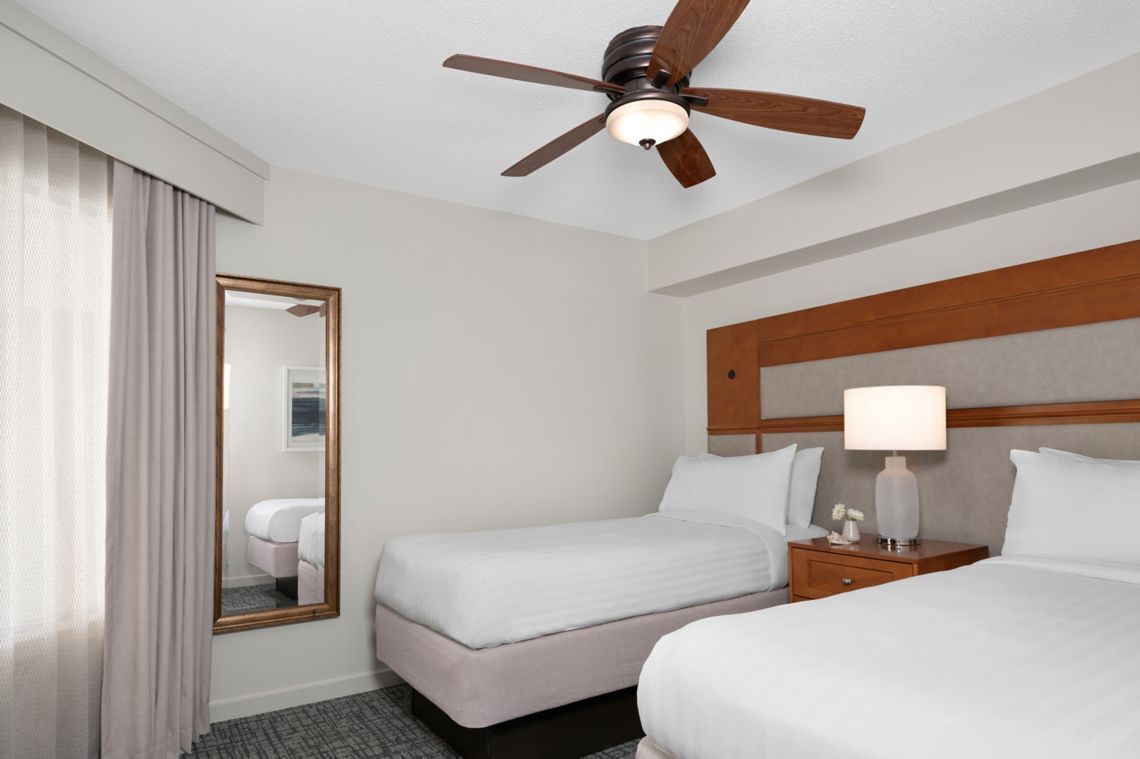 A hotel room with two beds, a ceiling fan, a nightstand with a lamp, and a large mirror.