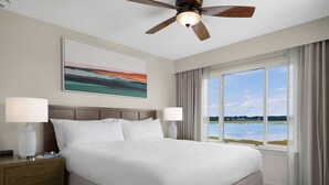 Ein Hotelzimmer mit Bett, zwei Nachttischlampen, Deckenventilator, Fenster mit Blick auf ein Gewässer und einem Bild an der Wand.