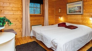 Quarto de cabana de madeira com uma cama, uma janela com cortinas, um abajur, um radiador e um quadro na parede.