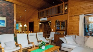 Interior de uma cabana de madeira com área de jantar, poltronas, um sofá e escada que leva a um andar superior.