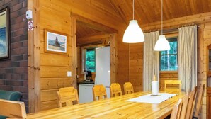 Interior em madeira com uma longa mesa de jantar, cadeiras, luminárias pendentes, área de cozinha e um quadro na parede.
