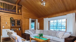 Interior de uma cabana de madeira com sofá, cadeiras, mesa e janela com vista para árvores.