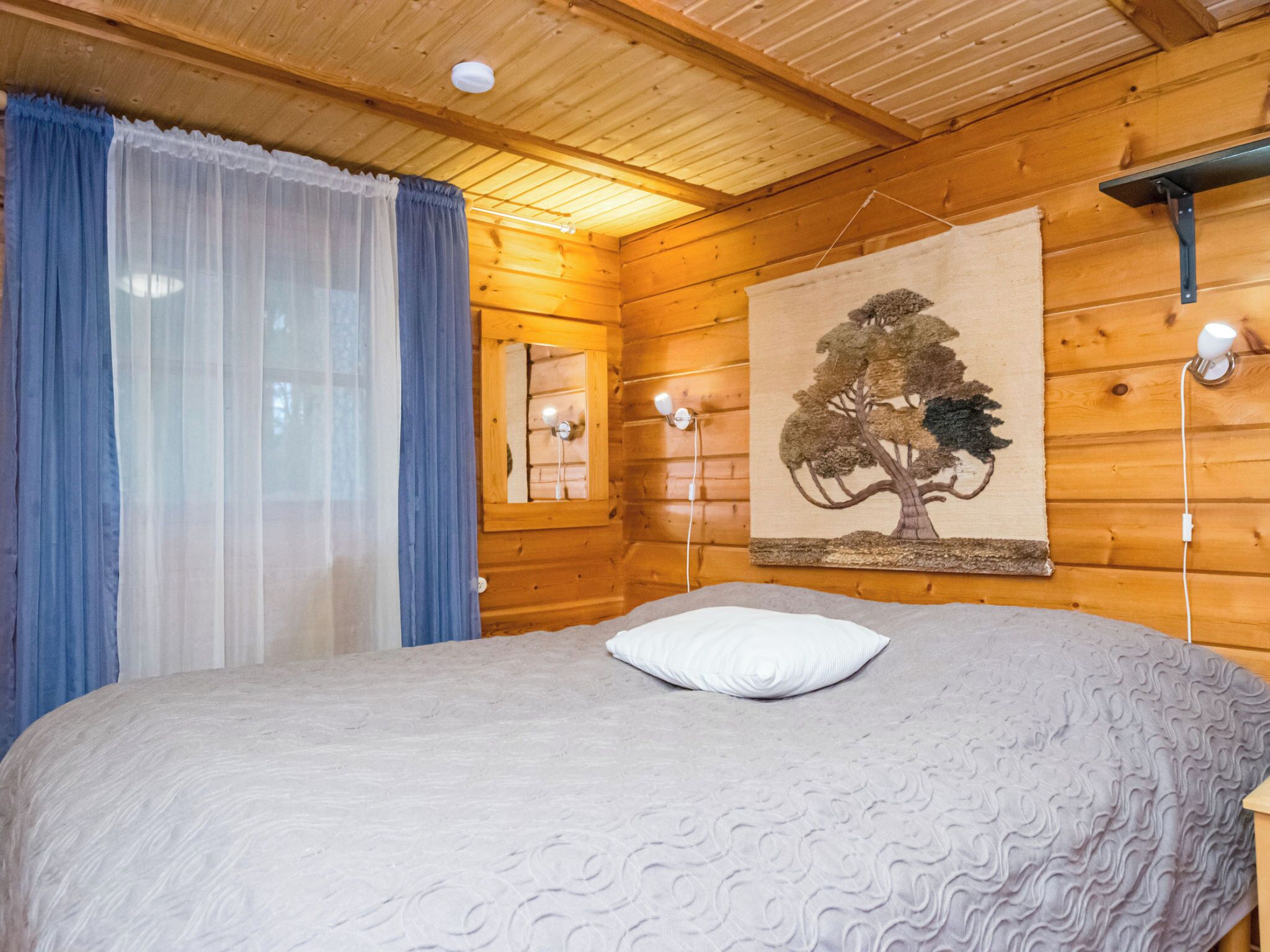 Ein Schlafzimmer einer Holzhütte mit einem Bett, einem Fenster mit Vorhängen, einer Wandlampe und einer Wandteppich.