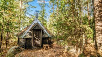 Mökki, jossa on maalaishenkinen tunnelma, kalteva katto, savupiippu ja puuovi, ja se sijaitsee metsän ympäröimänä.
