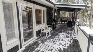 A covered outdoor deck with a grill, seating, and a view of a snowy forest.