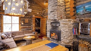 Interior de una cabaña de troncos con chimenea de piedra, muebles de madera y un televisor montado en la pared.