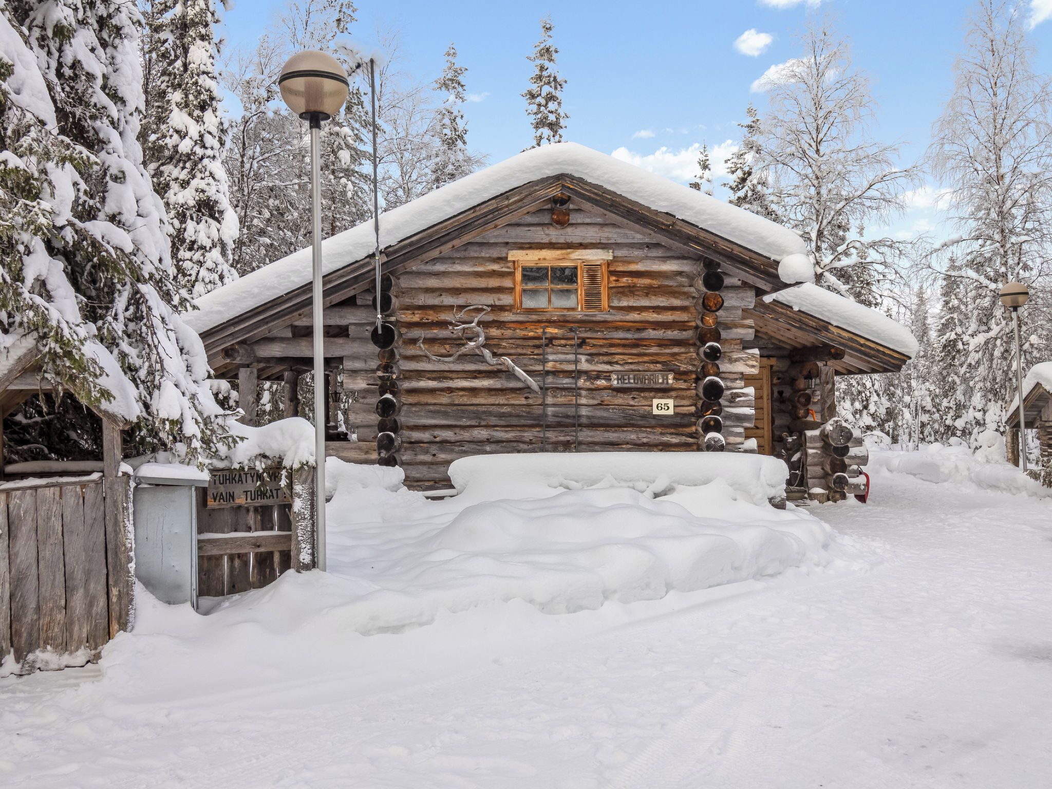 A log cabin with a snow-covered roof, a wooden porch, and a sign reading 'HELVARLIE 65'.
