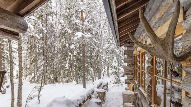 Una cabaña de troncos con exterior nevado, troncos de madera apilados afuera y una ventana con vistas a árboles cubiertos de nieve.
