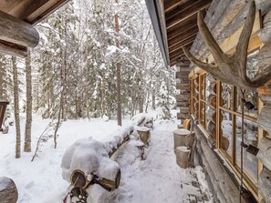 A log cabin with a snow-covered exterior, wooden logs stacked outside, and a window with a view of snow-laden trees. - Kelovarila by Interhome (Kuusamo)