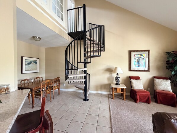 A spacious living area with a spiral staircase, a dining table with chairs, a small side table with a lamp, and a large plant.