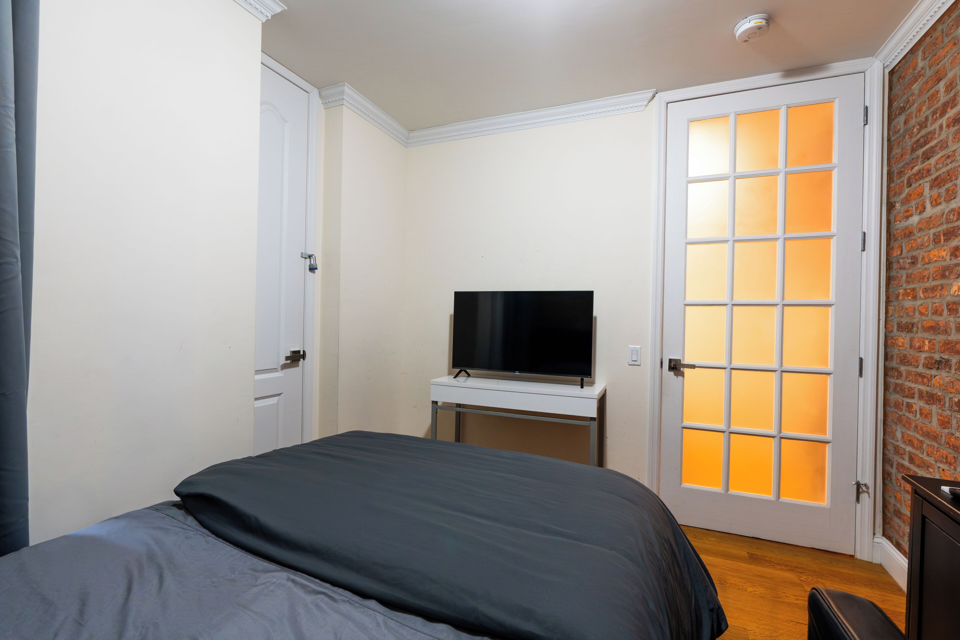 A bedroom with a bed, a television on a stand, a white door with frosted glass panels, and a brick wall.