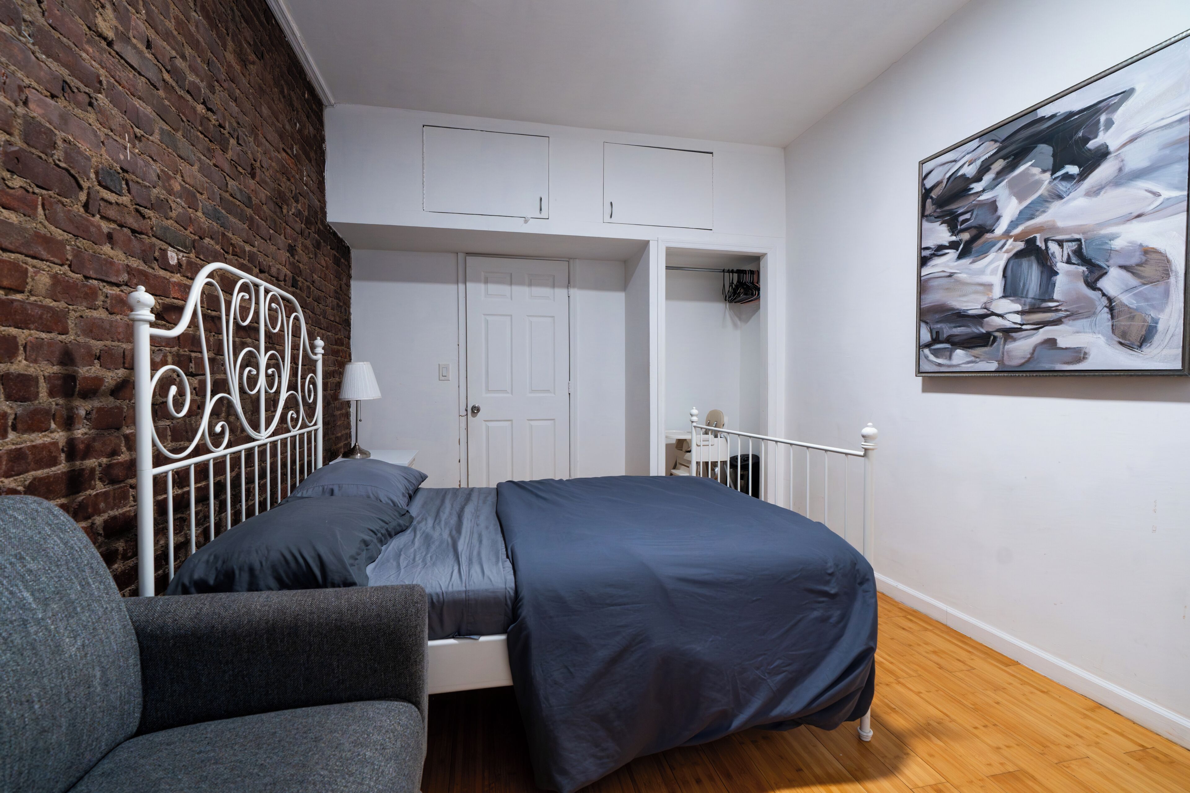 Un dormitorio con estructura de cama metálica, sofá gris, pared de ladrillo, puerta blanca y un cuadro en la pared.