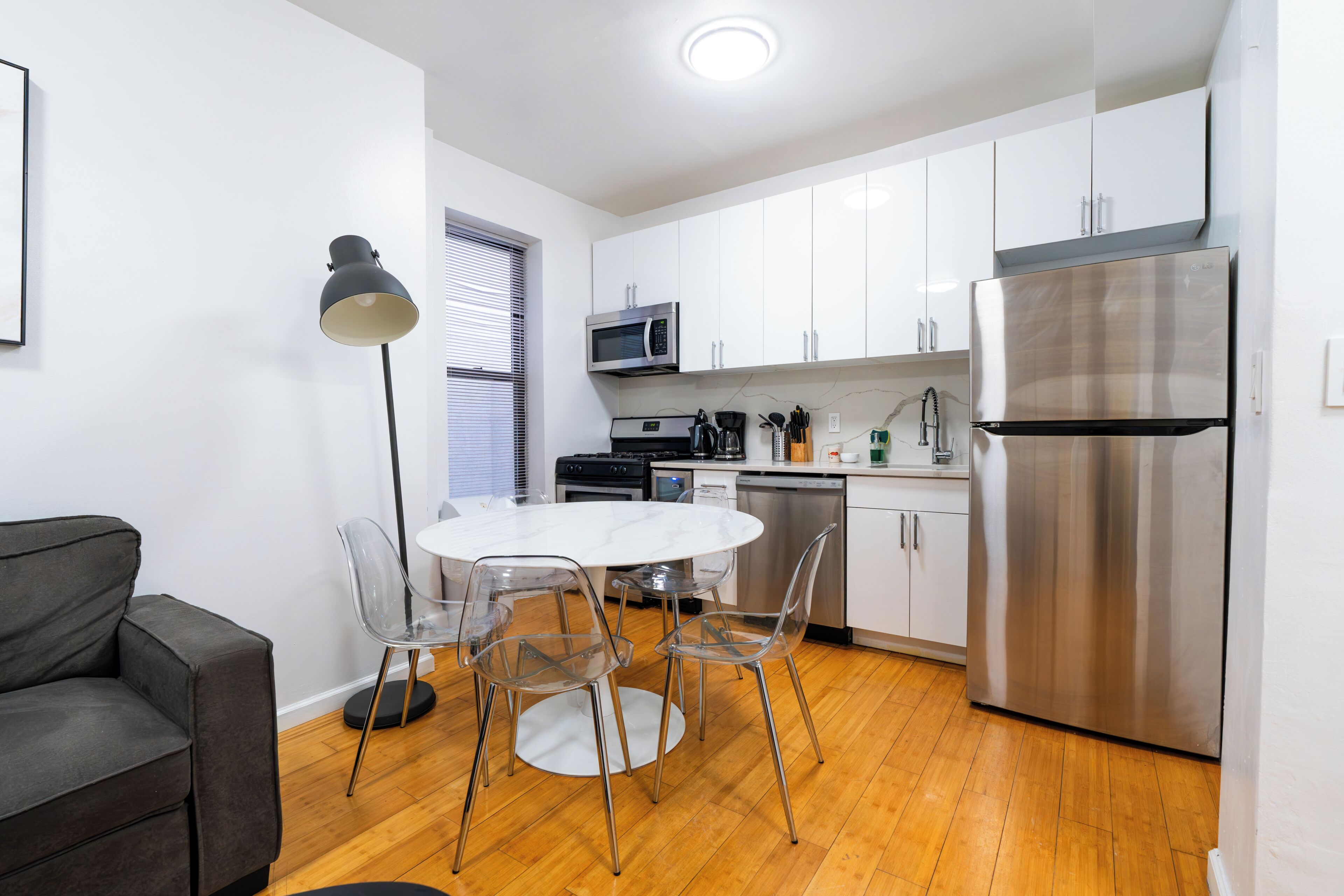 A modern kitchen with white cabinets, stainless steel appliances, a dining table with chairs, and a grey sofa.