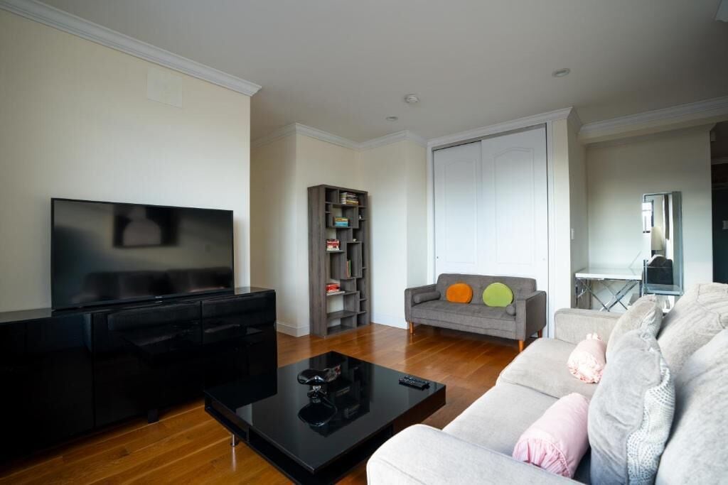 Una sala de estar con una televisión de pantalla plana, una mesa de centro negra, un sofá gris con cojines, una estantería y una puerta.