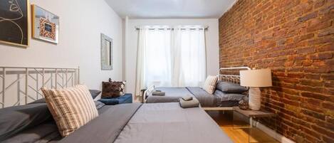 A room with two beds, a brick wall, a window with curtains, and a lamp on a side table.