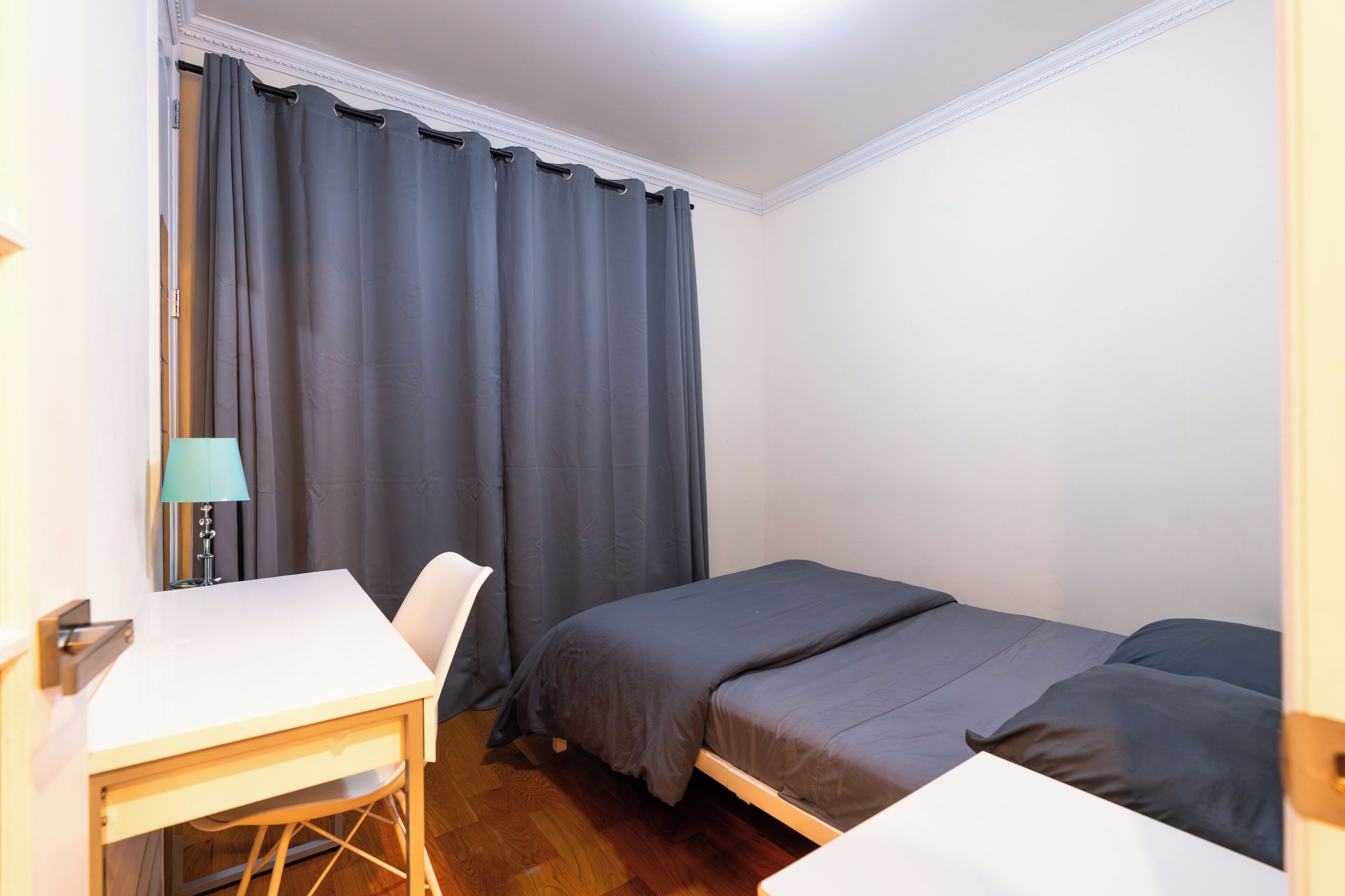 Ein kleines Zimmer mit einem Einzelbett, einem Schreibtisch, einem Stuhl, einer Lampe und einem Fenster mit Vorhängen.