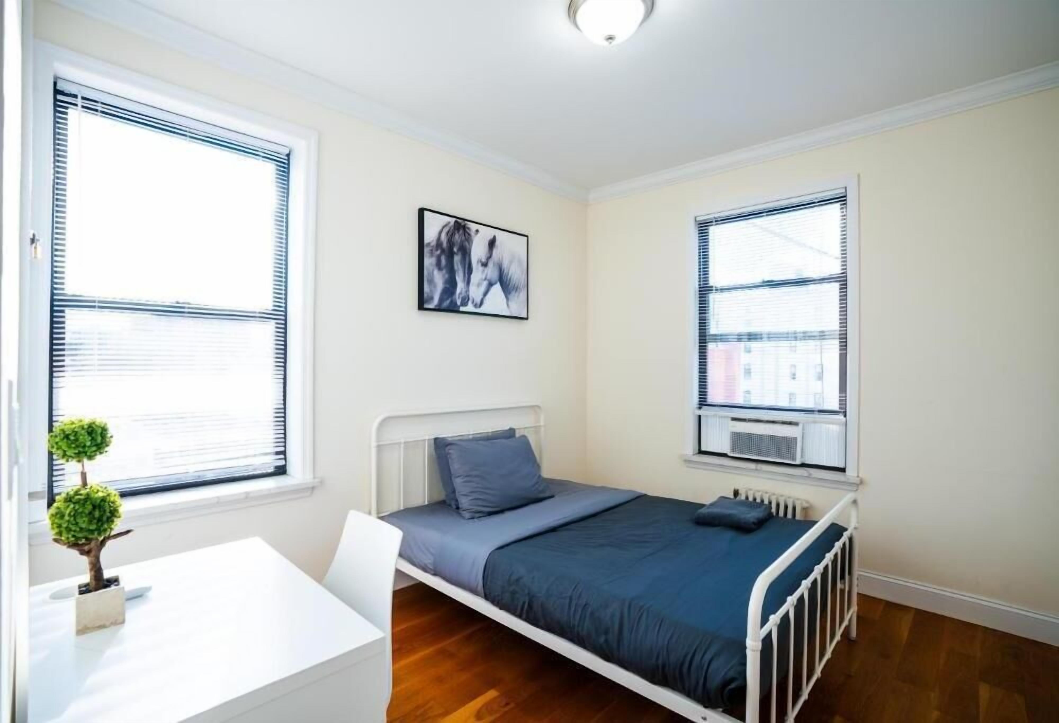 A bedroom with a bed, a desk, a chair, a radiator, and a window with blinds.