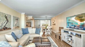 A living room with a sectional sofa, a dining area with a table and chairs, a TV on a stand, and artwork on the walls. - 2 Bedroom beach/pool view,37ft balcony, boat slip,6 month rental only, May-Oct. (Marco Island)
