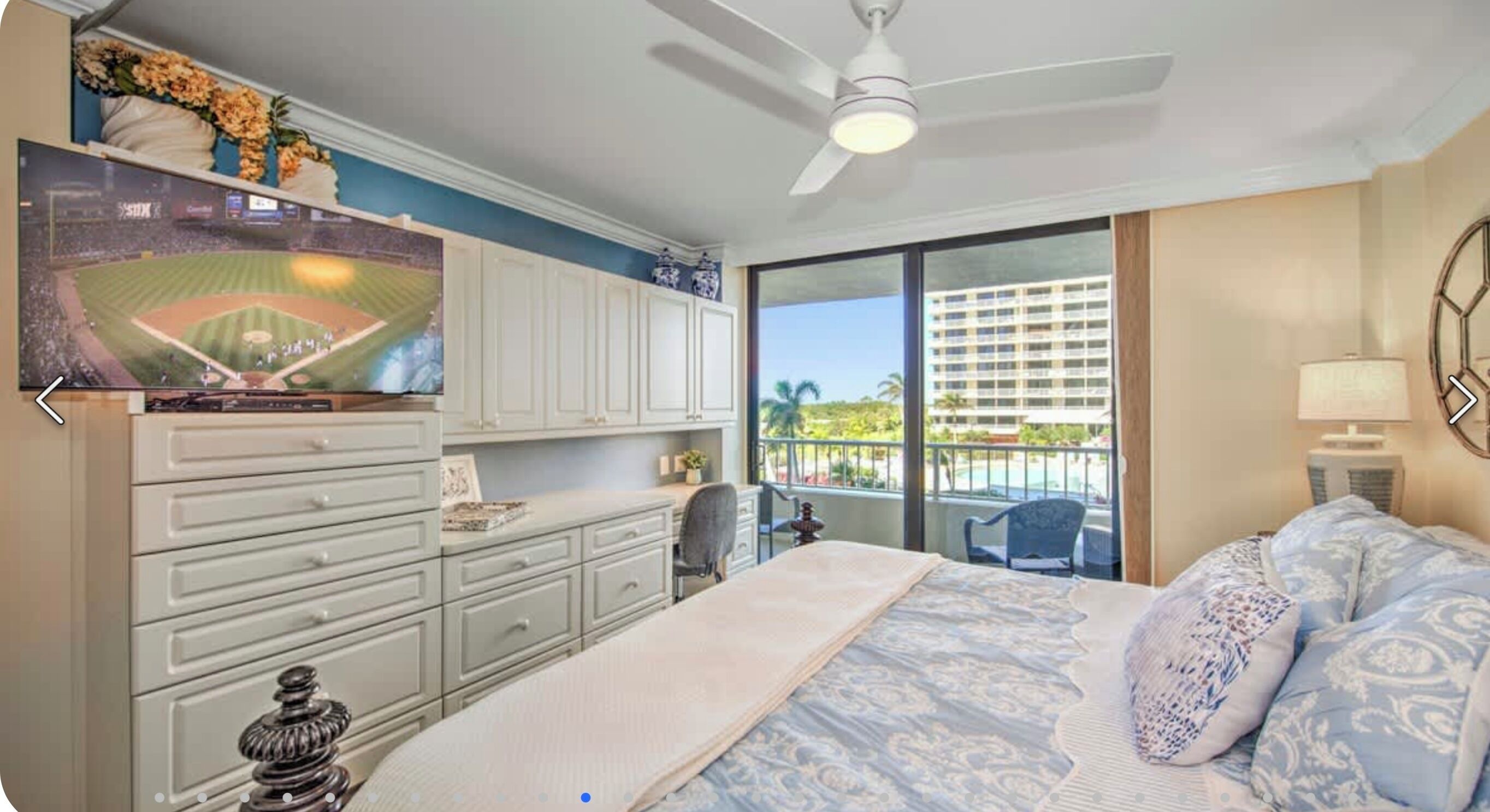 A bedroom with a bed, a desk, a chair, a ceiling fan, and a view of the outdoors.