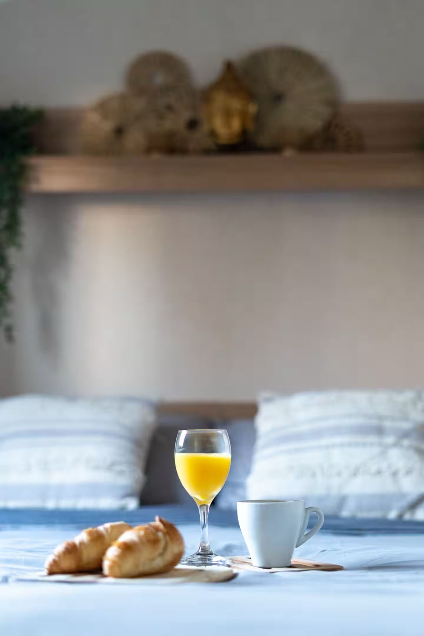 Un lit avec une couette blanche, un verre de jus d’orange, deux croissants et une tasse blanche sur un plateau en bois.