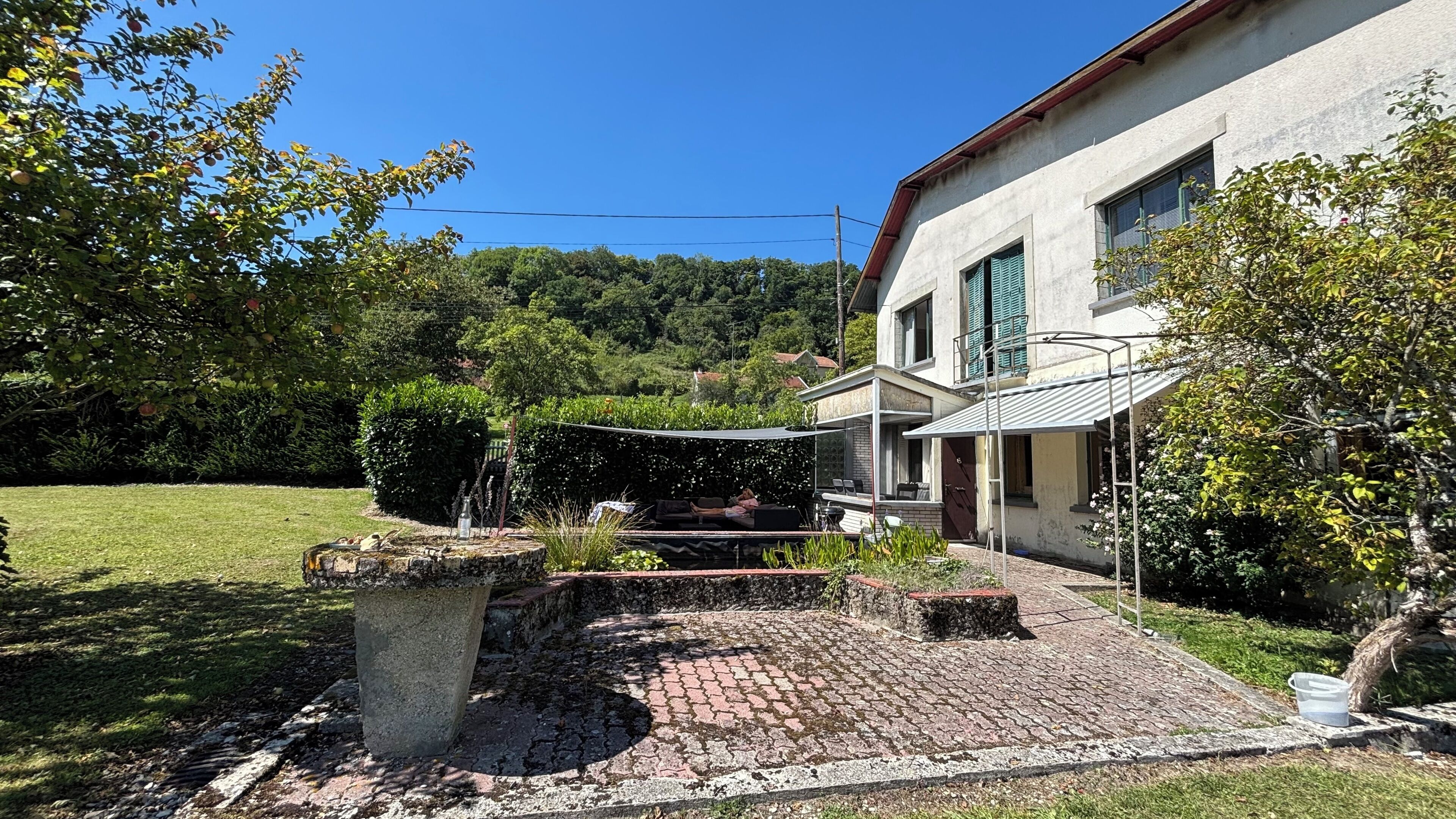 Ein Haus mit gepflastertem Innenhof, einer überdachten Sitzgelegenheit und einem Garten mit Bäumen und Sträuchern.