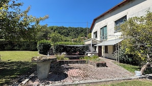 A house with a paved courtyard, a covered outdoor seating area, and a garden with trees and shrubs.