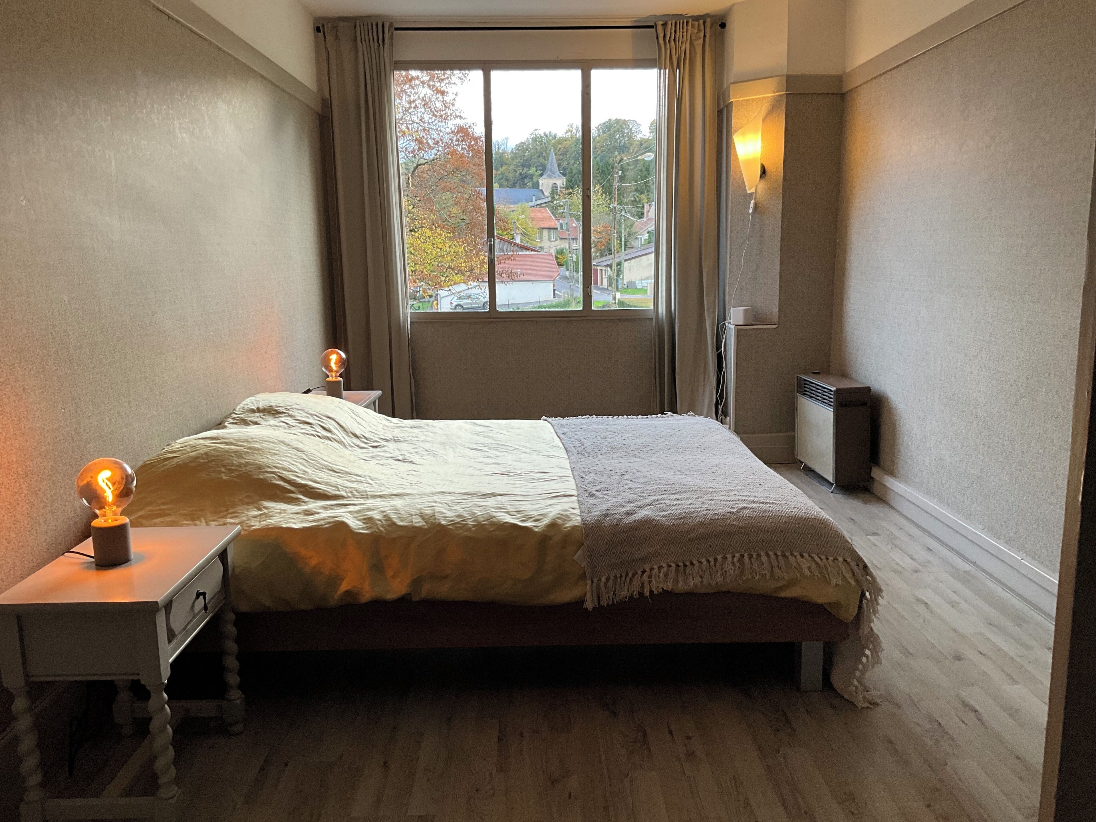 Ein Schlafzimmer mit einem Bett, einem Nachttisch, einer Lampe, einem Fenster und einer Wandheizung.