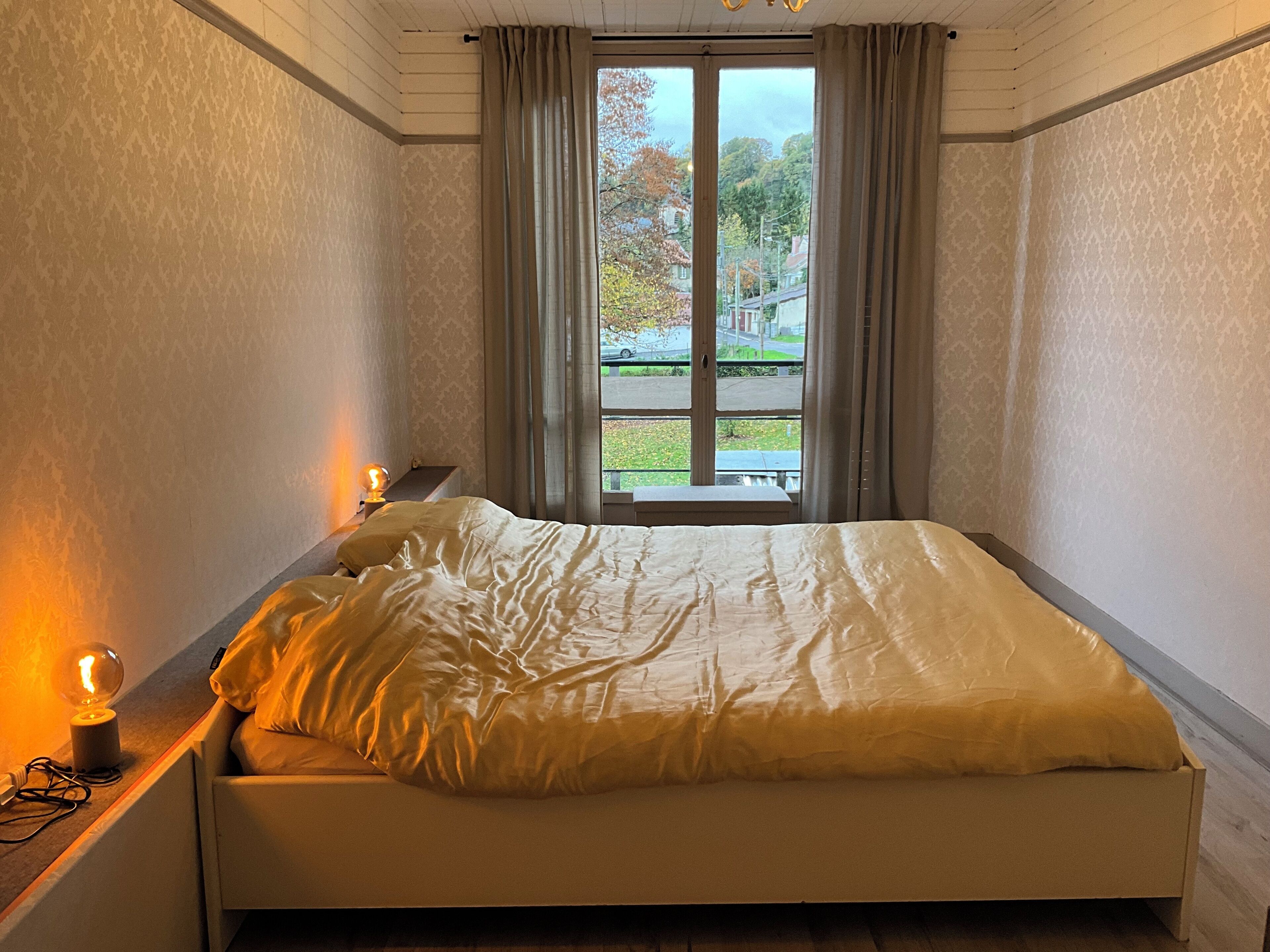Ein Schlafzimmer mit Bett, Nachttisch, Lampe und Fenster mit Blick nach draußen.