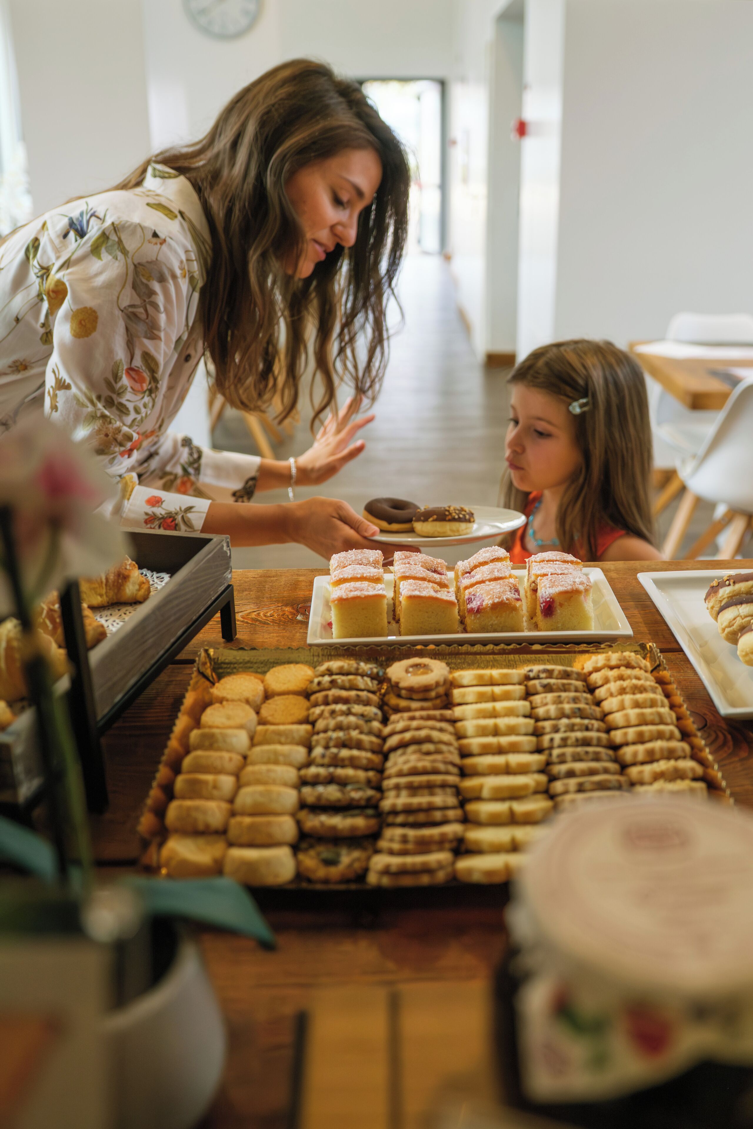 A woman serving food to a child at a table with various pastries and cookies.