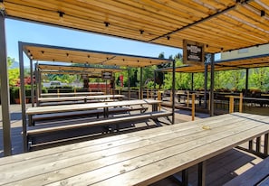 An outdoor seating area with wooden tables and benches, covered by a wooden pergola, and signs indicating a restaurant.