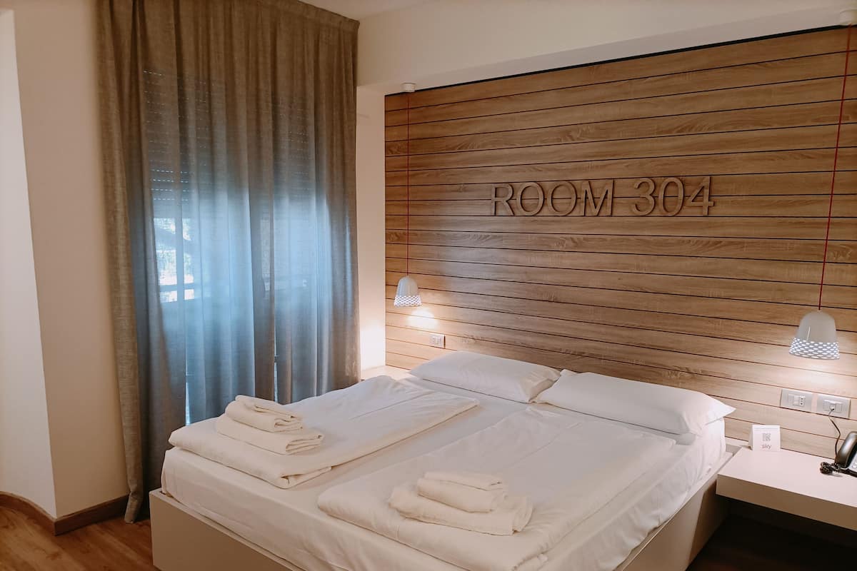 A hotel room with a wooden headboard, a bed with white linens, a bedside table with a lamp, and a window with curtains.