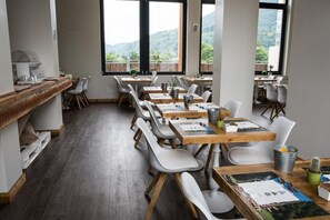 A dining area with tables and chairs, a wooden counter, and large windows with a view of greenery.