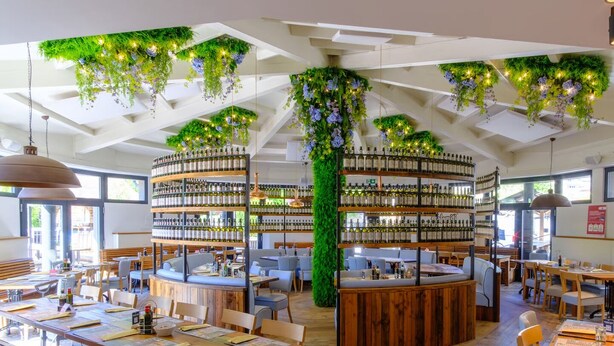 Een restaurant met houten tafels en stoelen, en een bar in het midden met flessenkasten.