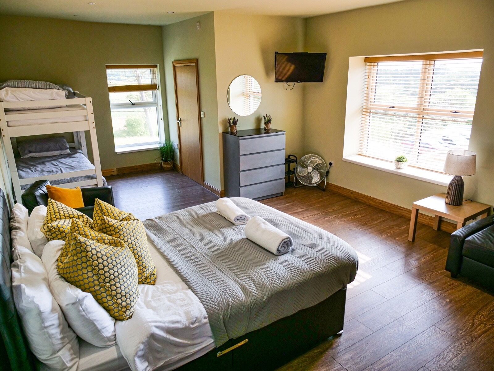 Un dormitorio con una cama individual superpuesta, una cama individual con toallas enrolladas, un aparador, un ventilador, un televisor y una ventana con persianas.