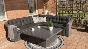 A black leather sectional sofa with a matching coffee table, a potted plant, and a wicker chair on a patio with a brick wall and a wooden fence. - 4 Star Holiday Home in Skaret (Nyhamnslage)