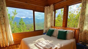 Une chambre située dans une cabane en bois, équipée d’un lit, d’un ventilateur de plafond, d’un appareil de climatisation et de grandes fenêtres offrant une vue sur la montagne.