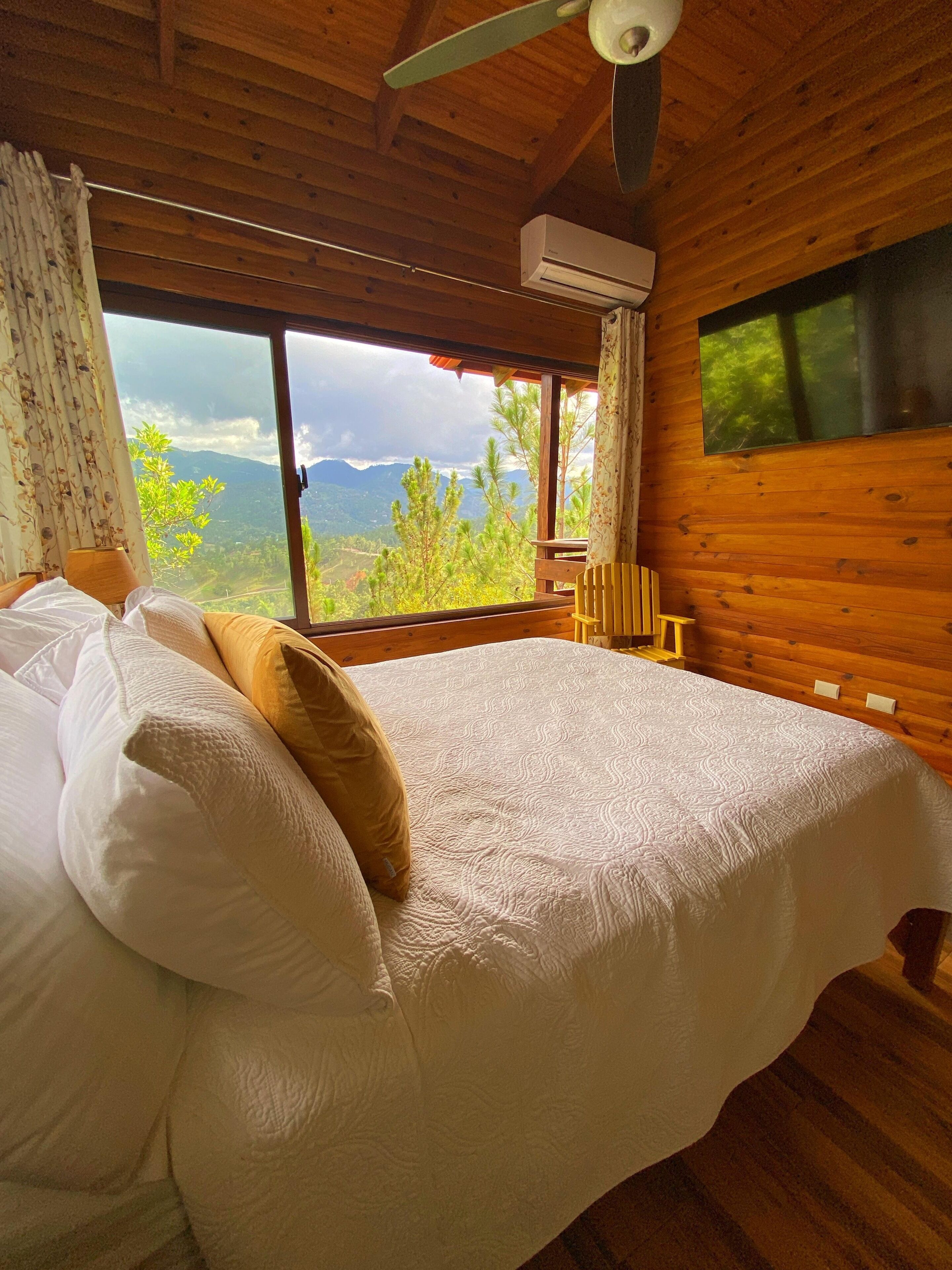 Une chambre dans un chalet en bois, avec un lit, des oreillers, une fenêtre donnant sur les montagnes et un appareil de climatisation.