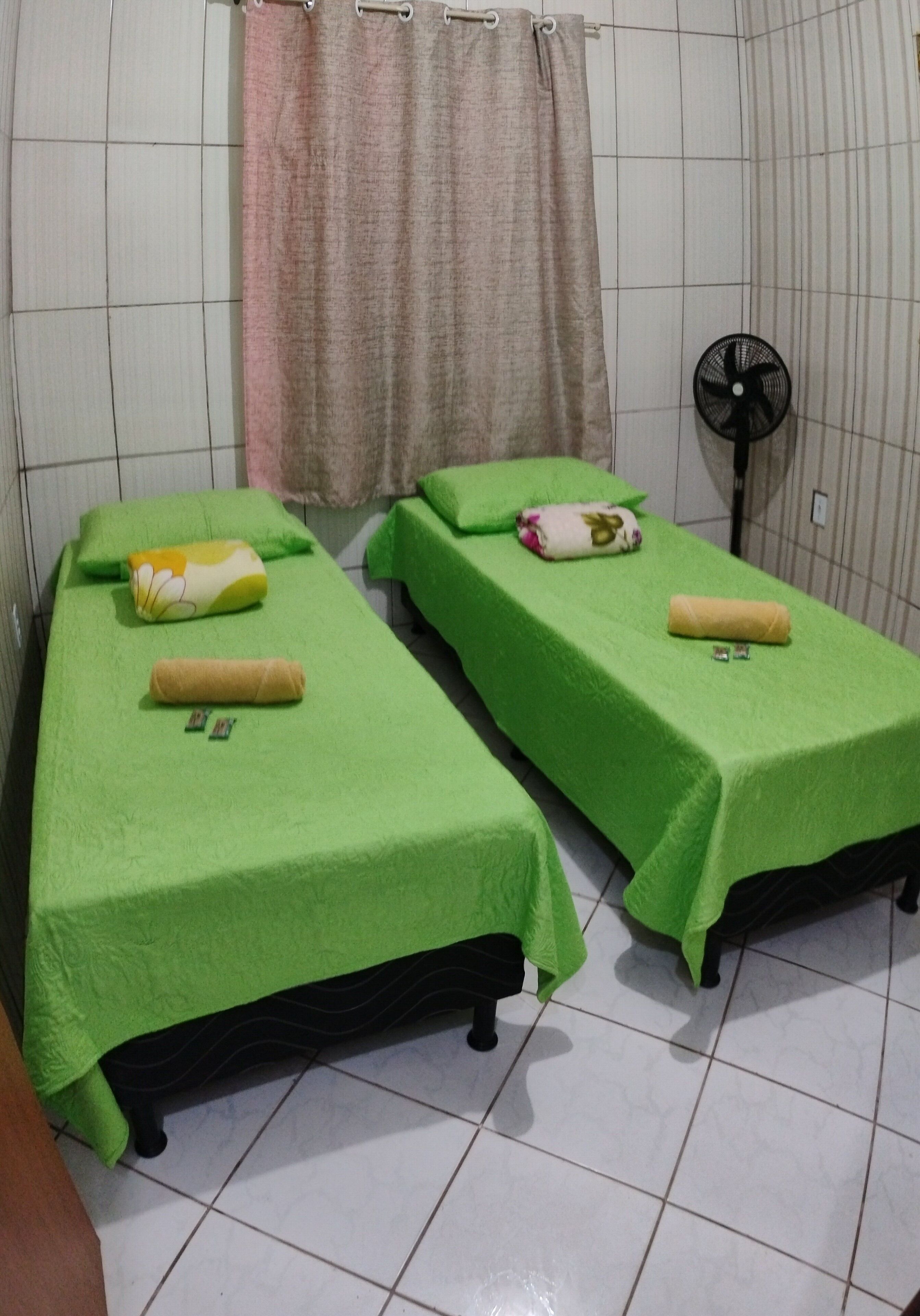Una stanza con due letti singoli, ognuno con una coperta verde e un cuscino, una ventola e una tenda.