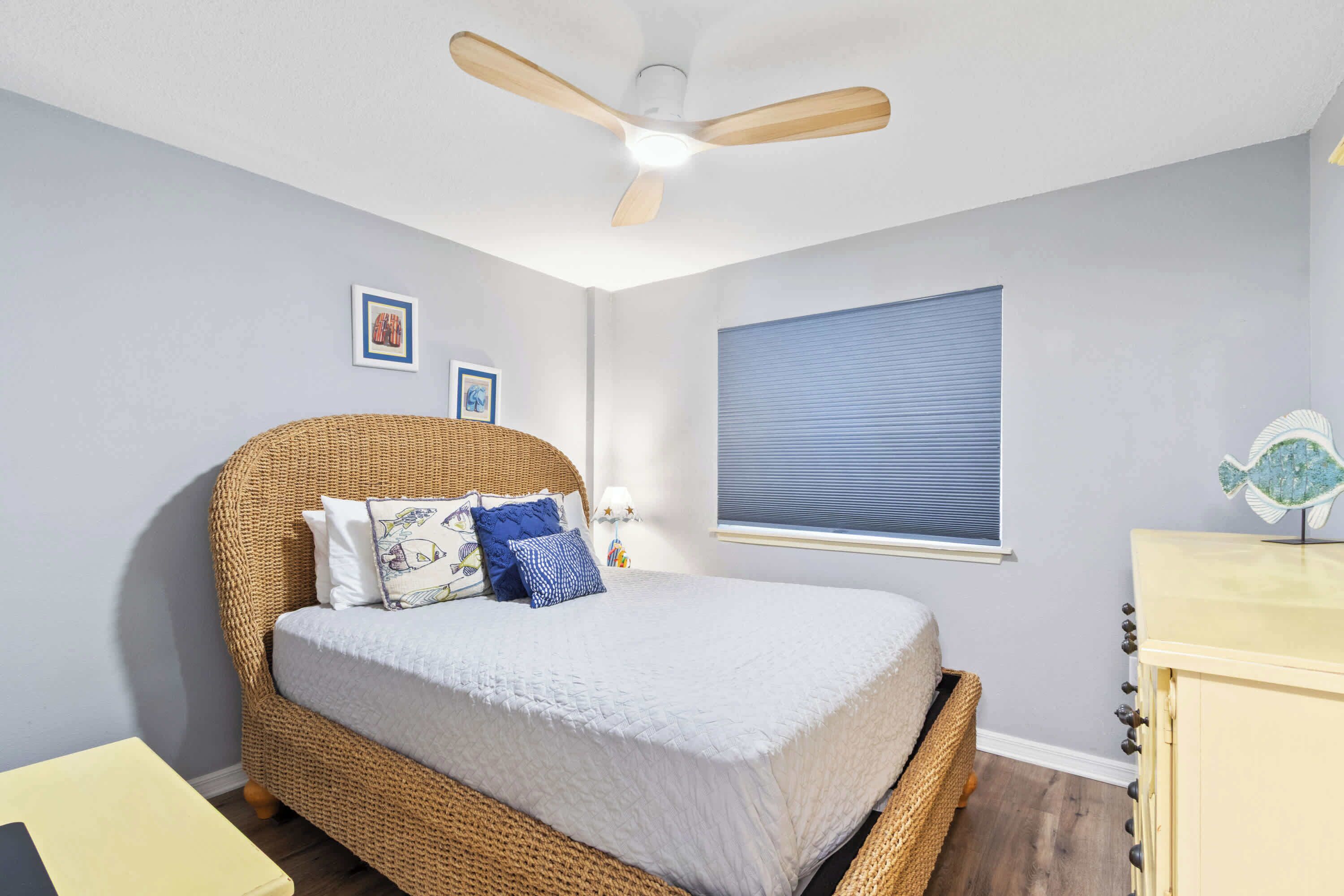 Quarto com cama de vime, ventoinha no teto, janela com persianas e cómoda com um objeto decorativo.