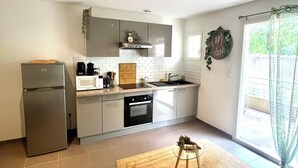 A modern kitchen with a refrigerator, oven, and sink. There is a wooden table with a rug, and a window with a view of greenery. - Cosy Corner (Villeneuve-Loubet)