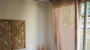 A bedroom with a bed, a bedside table, a wall-mounted air conditioner, and a window with a curtain. - Cosy Corner (Villeneuve-Loubet)