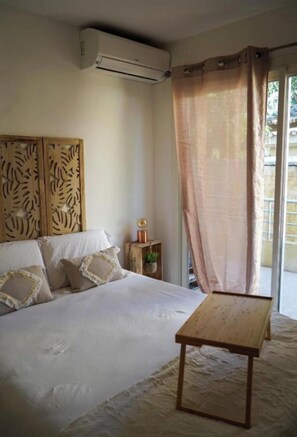 A bedroom with a bed, a bedside table, a wall-mounted air conditioner, and a window with a curtain. - Cosy Corner (Villeneuve-Loubet)