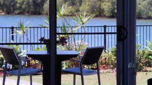 Une terrasse avec une table et des chaises, une clôture en métal et une vue sur un lac bordé d’arbres.