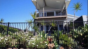 Une maison moderne à deux étages, dotée d’un balcon, entourée d’une clôture en métal noir et agrémentée de diverses plantes tropicales.