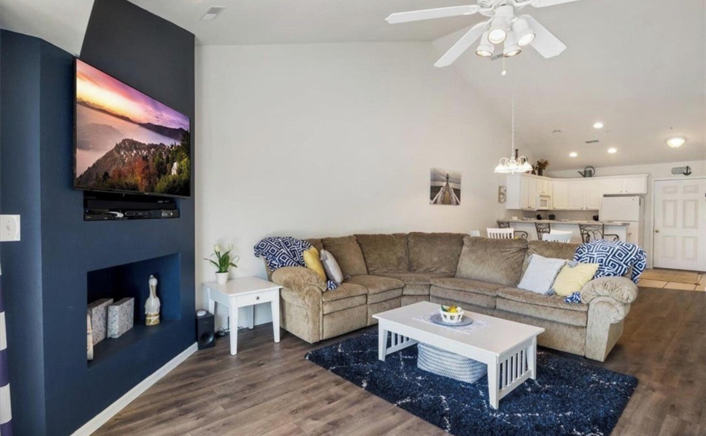 A modern living room with a sectional sofa, a flat-screen TV, a ceiling fan, and a kitchen area in the background.