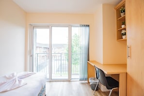 Une chambre d’hôtel comprenant un lit, un bureau, une chaise, une poubelle et une étagère en bois ornée de plantes.